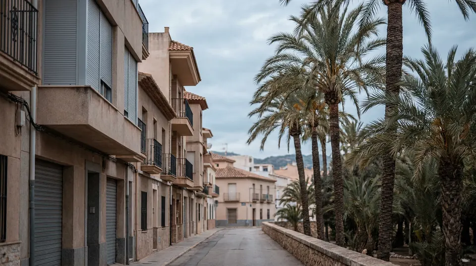 Calle de bloques de pisos con persianas enrollables junto a un huerto de palmeras